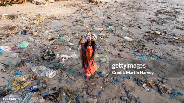 aerial. black woman selling fish walks with a basket on her head through the dreadful plastic pollution that lines the water's edge of the fish market in hann, senegal - ugly black women ストックフォトと画像