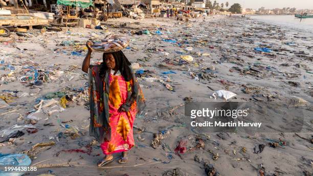 aerial. black woman selling fish walks with a basket on her head through the dreadful plastic pollution that lines the water's edge of the fish market in hann, senegal - ugly black women ストックフォトと画像