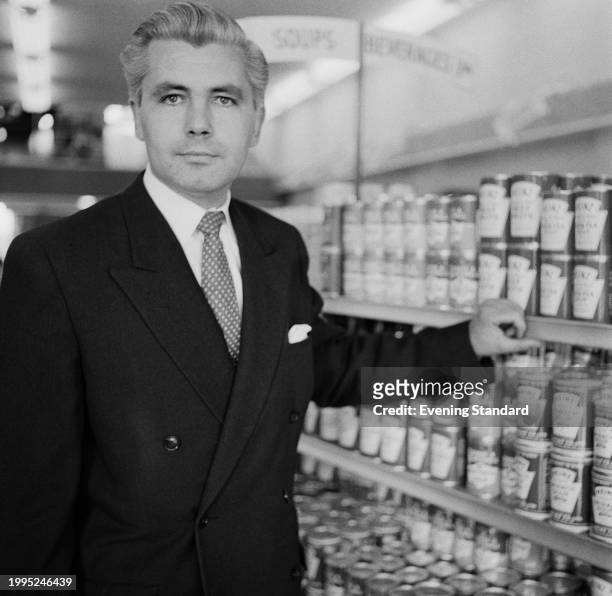 Premier Supermarkets head, Patrick Galvani leaning on shelves of tinned food in a supermarket aisle, Britain, July 25th 1957.