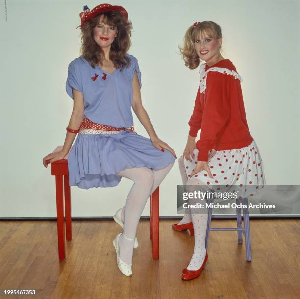 One woman models a blue short-sleeve dress with a red polka dot belt and a red hat sits on a red table, sitting beside a woman modelling a red top...