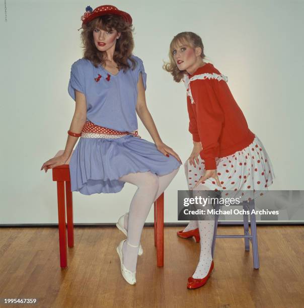 One woman models a blue short-sleeve dress with a red polka dot belt and a red hat sits on a red table, sitting beside a woman modelling a red top...