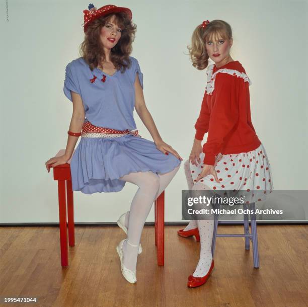 One woman models a blue short-sleeve dress with a red polka dot belt and a red hat sits on a red table, sitting beside a woman modelling a red top...