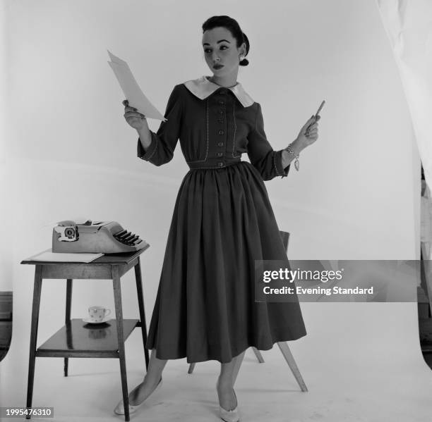 Studio shot of a fashion model wearing an office dress while holding a sheet of paper beside a typewriter on a side table, December 11th 1956.