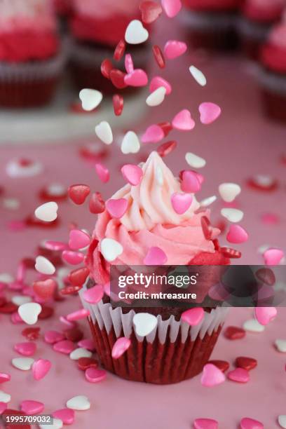 close-up image homemade, red velvet cupcake in paper cake case being sprinkled with red, pink and white candy hearts, decorated with ombre effect pink butter icing piped swirl, sweets floating mid-air against pink background, valentine's day and romance - cake case stock pictures, royalty-free photos & images