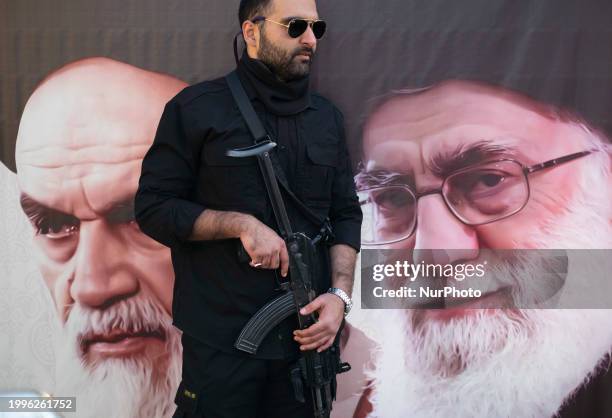 An Iranian armed military personnel is monitoring an area while standing guard in front of a banner featuring portraits of Iran's Supreme Leader...