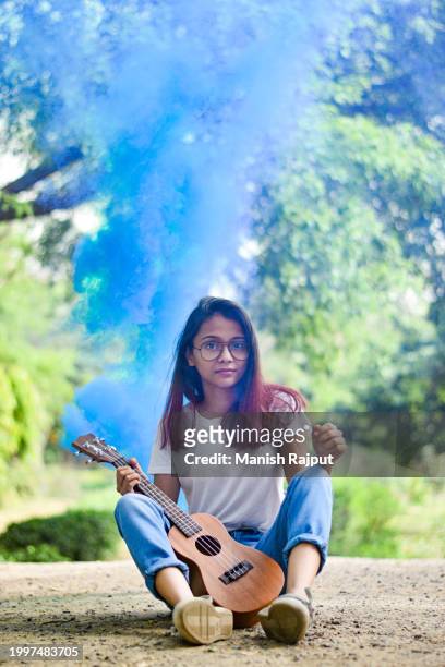 a young musician sits in the garden, holding a ukulele, with blue smoke behind her. - blue acoustic guitar stock pictures, royalty-free photos & images