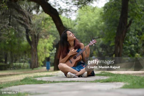 a young female musician, dressed in a blue floral dress, sits in the garden playing the ukulele. - blue acoustic guitar stock pictures, royalty-free photos & images