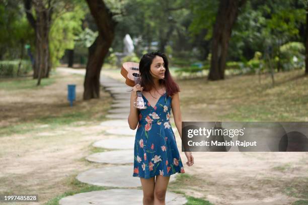 a young female musician, dressed in a blue floral dress, stands in the garden holding the ukulele on shoulder. - blue acoustic guitar stock pictures, royalty-free photos & images
