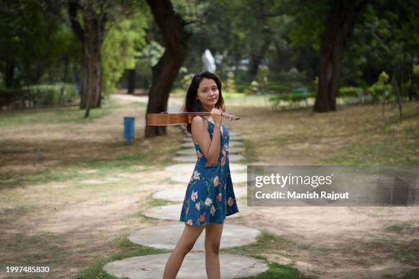 a young female musician, dressed in a blue floral dress, stands in the garden holding the ukulele on shoulder. - blue acoustic guitar stock pictures, royalty-free photos & images