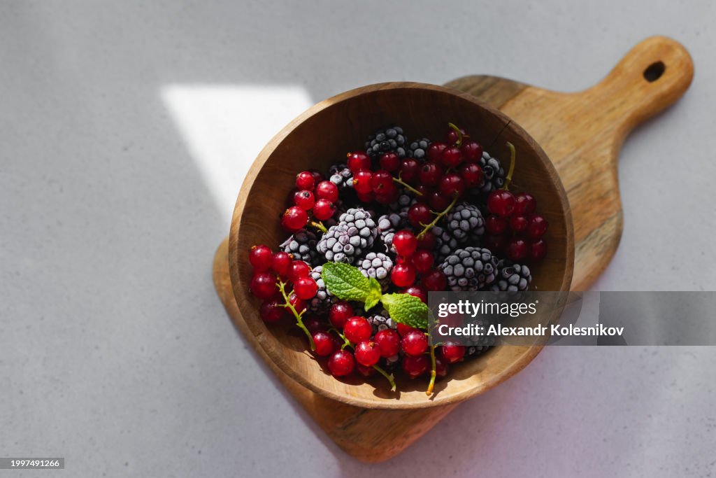 Wooden bowl with frozen blackberry and red currant on light background with copy space. Summer berry food. Table top view