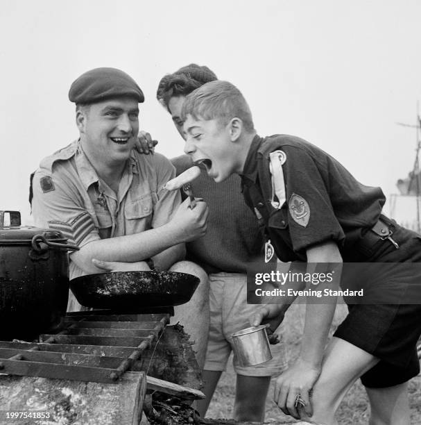 Boy Scout Richard Heyo opens his mouth to eat a sausage offered by an army sergeant during the 9th World Scout Jamboree, Sutton Park, Sutton...