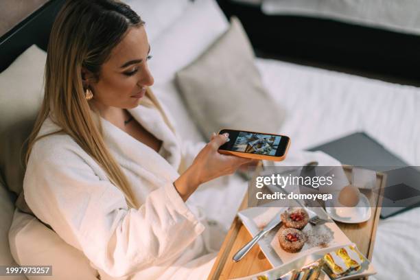 mujer joven tomando foto de un desayuno de servicio a la habitación en la cama - servicio de habitaciones fotografías e imágenes de stock