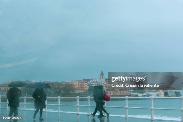 silhouettes of people with umbrellas walking along the promenade of the city of gijón on a rainy winter day. - gijón fotografías e imágenes de stock