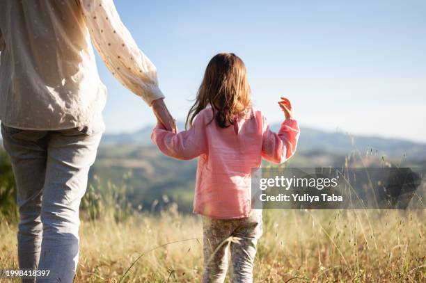 vista trasera de una madre sosteniendo la mano de su hija en un prado y mirando a la vista. - padre e hijas fotografías e imágenes de stock