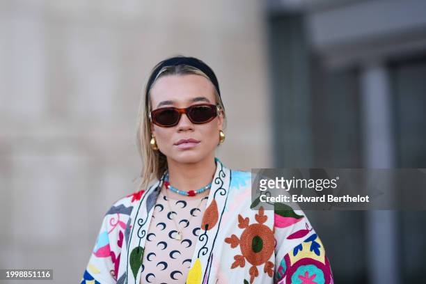 Guest wears a headband, golden earrings , a colored floral print coat , a blue and red necklace, a Marine Serre top with printed monograms, outside...