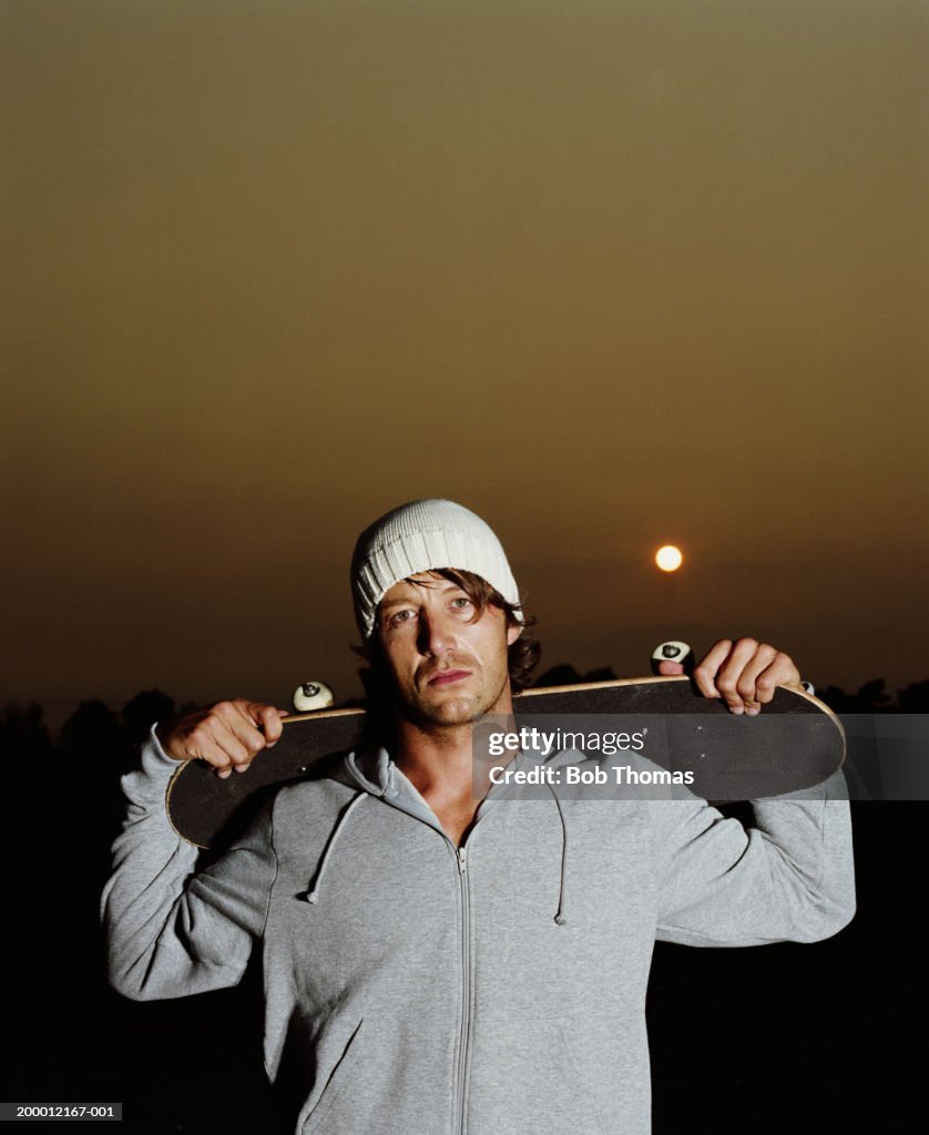 Young man holding skateboard behind head, sunset, portrait