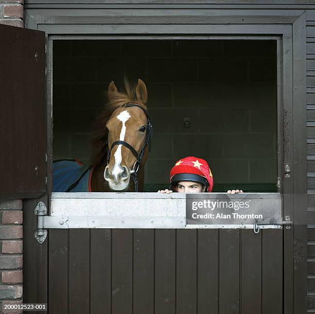 teenage jockey (15-17) peering over stable door next to horse - horse jockey stock-fotos und bilder