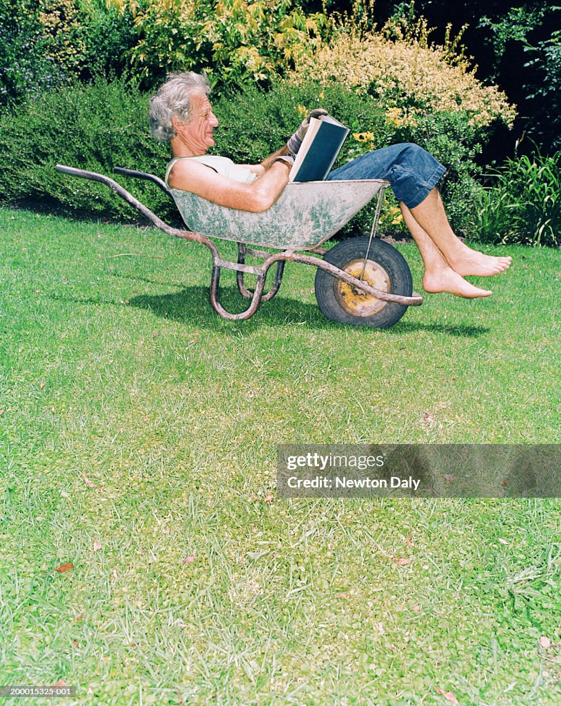 Mature man relaxing in wheelbarrow reading book, profile