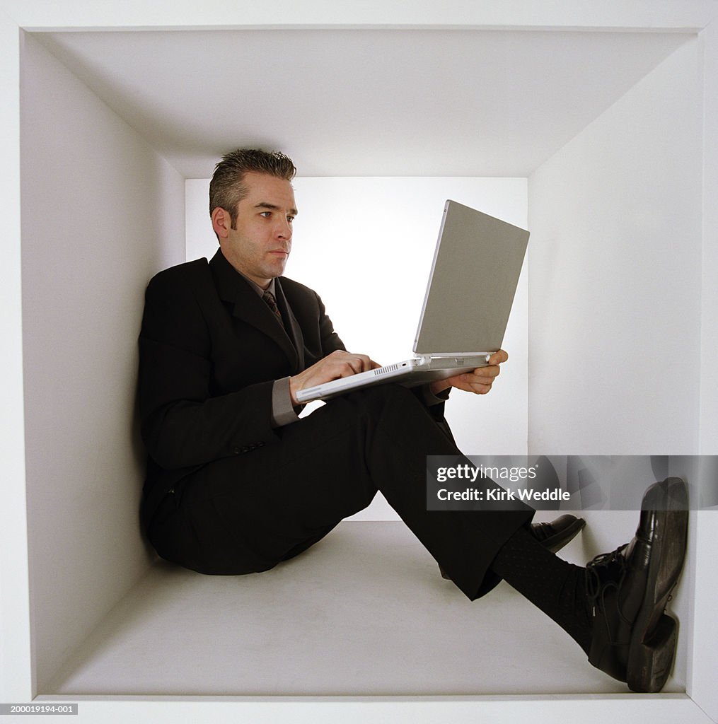Man sitting inside white box, working on laptop computer