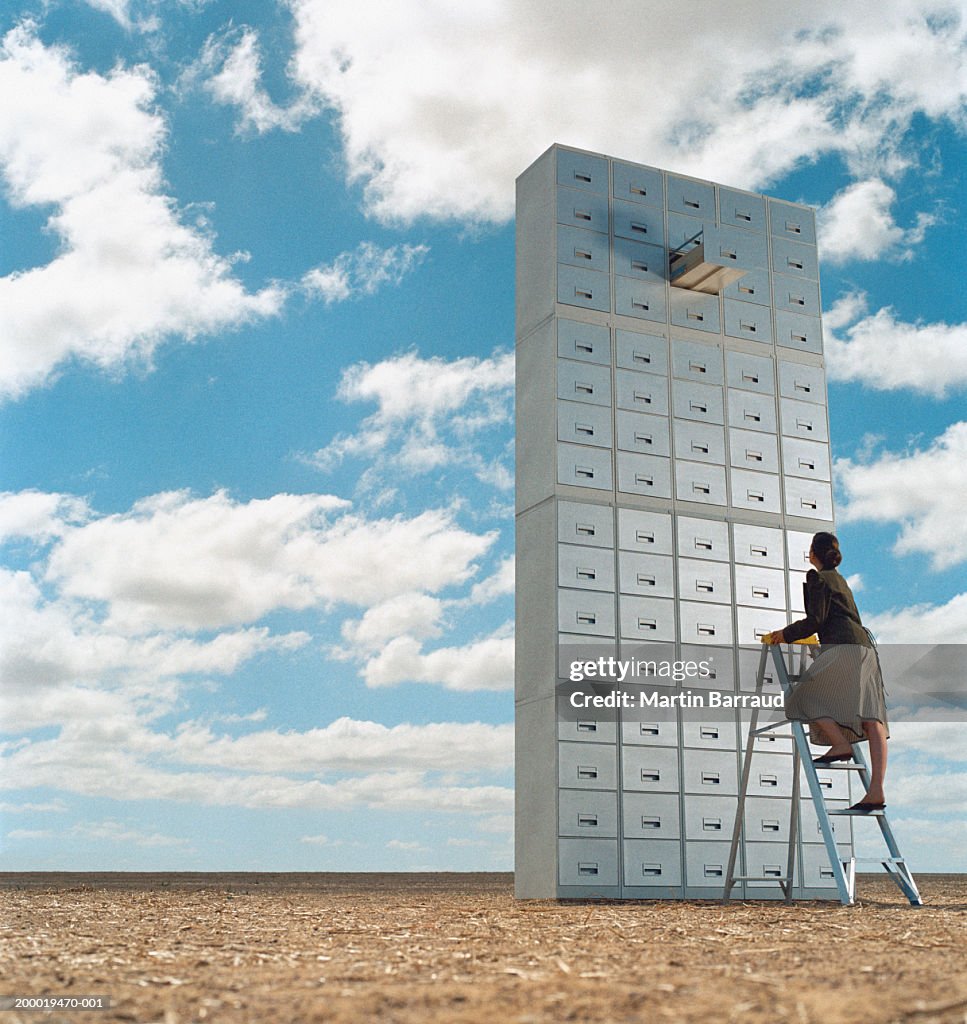 Woman climbing ladder towards open drawer in tower of filing cabinets