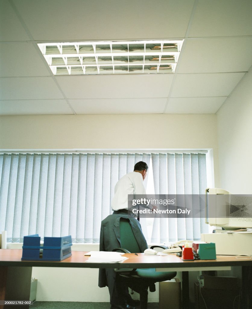 Mature businessman peering through blinds in office, rear view