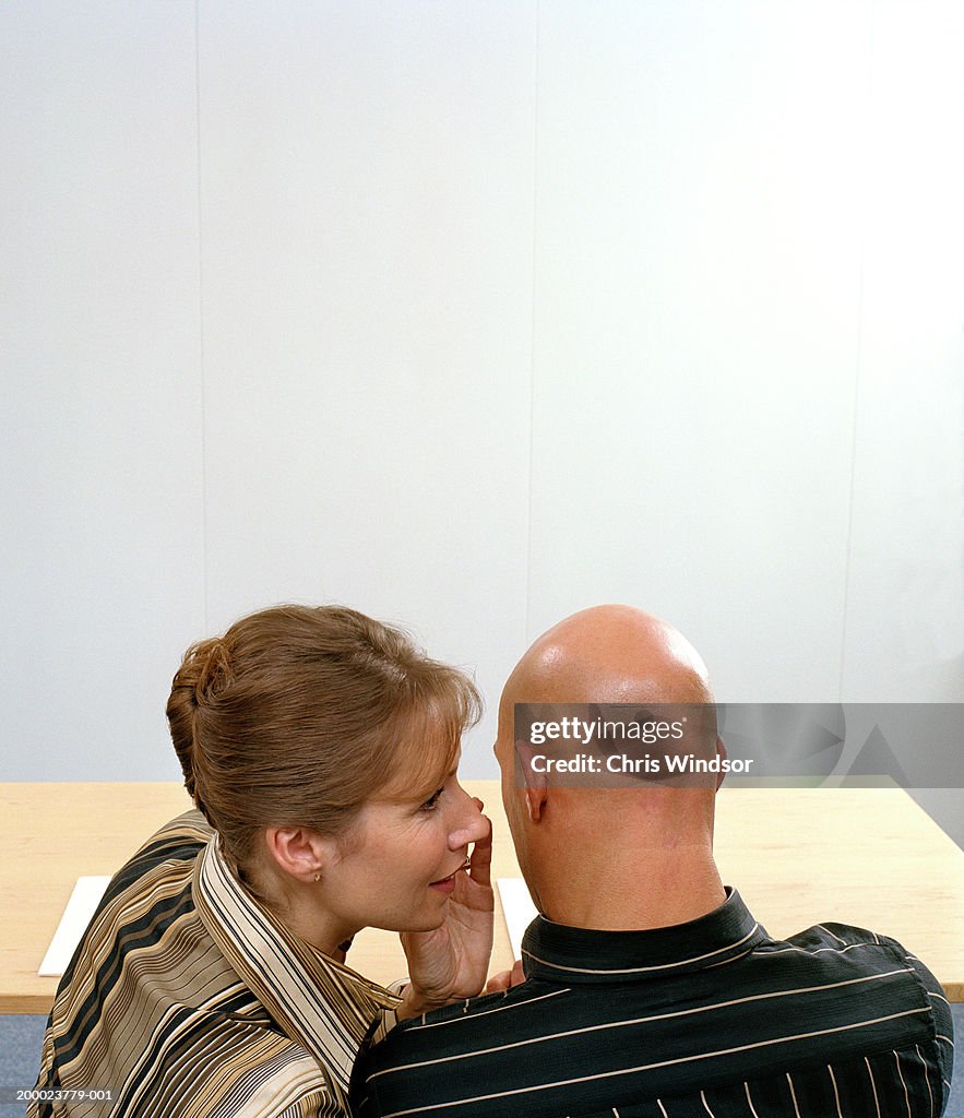 Businessman and woman at desk, woman whispering to man, rear view