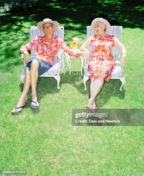 mature couple relaxing on garden chairs, holding hands, smiling - hawaiian shirt stock-fotos und bilder