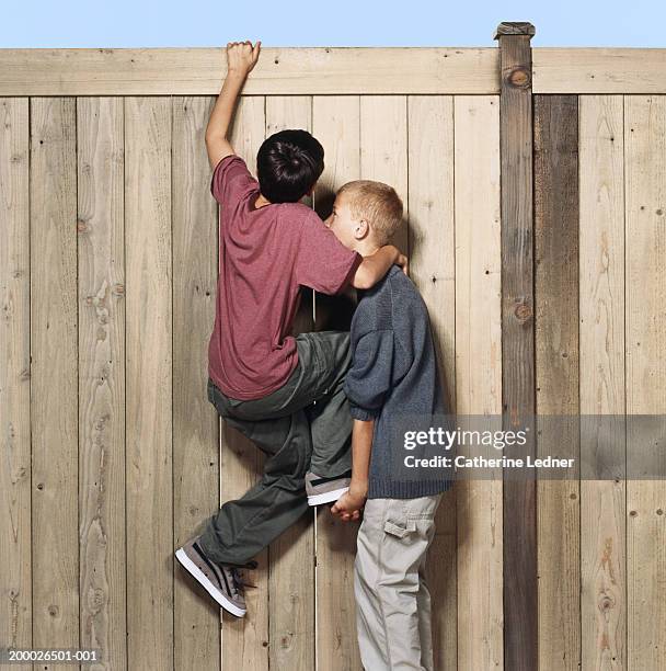 two boys (10-12) cimbing over fence, one boy lifting the other - one friend helping two other imagens e fotografias de stock