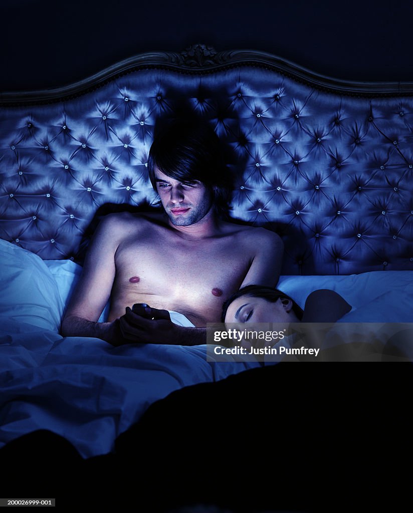 Young couple in bed, woman asleep, man using mobile phone, night