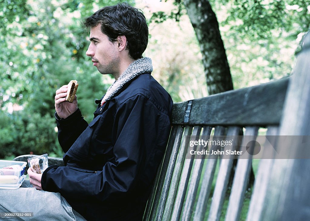 Young man eating sandwich on park bench, profile