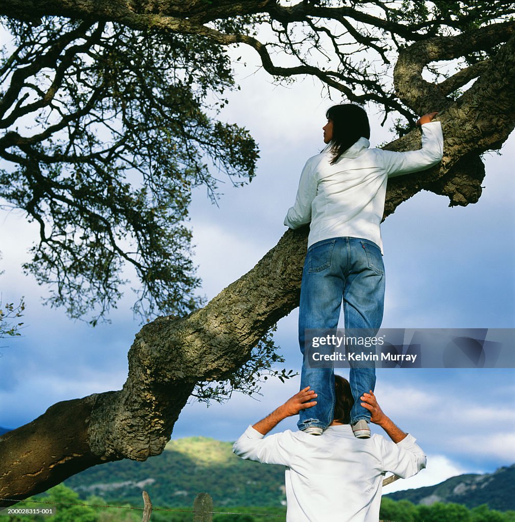 Woman standing on man's shoulders, holding onto tree branch