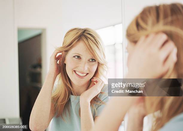 young woman adjusting hair in mirror, smiling - woman mirror stockfoto's en -beelden