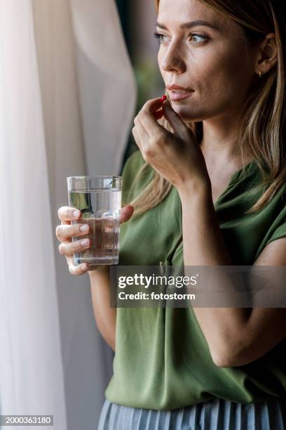 femme debout près de la fenêtre, prenant ses médicaments - comprimés photos et images de collection