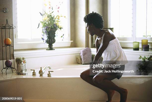 young woman sitting on edge of bathtub - african american woman bath stock pictures, royalty-free photos & images