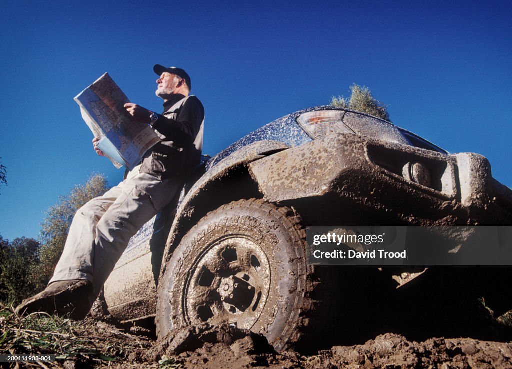 Mature man sitting on dirty car, holding map, low angle view