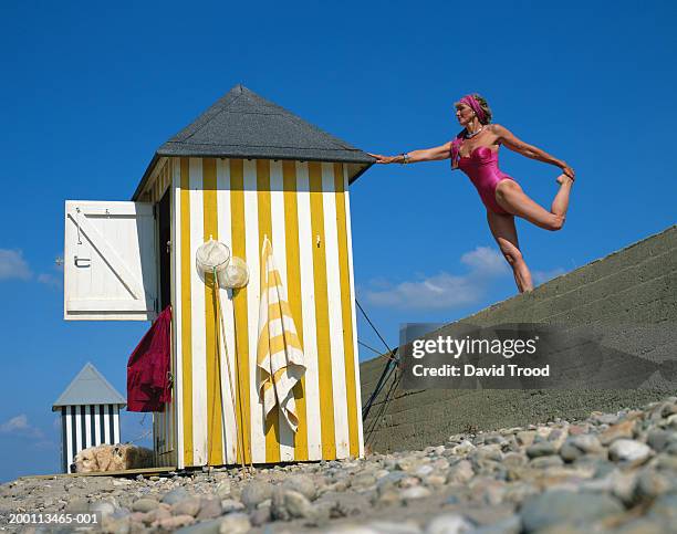 mature woman standing on one leg on wall, touching roof of beach hut - animal leg stockfoto's en -beelden