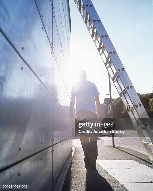 young man walking along street, ladder in foreground, rear view - ladder stock pictures, royalty-free photos & images