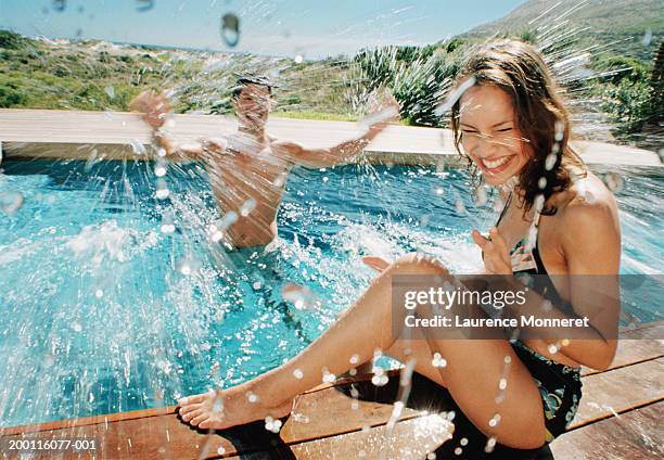 young couple, man in pool splashing woman sitting at edge - women by pool stock-fotos und bilder