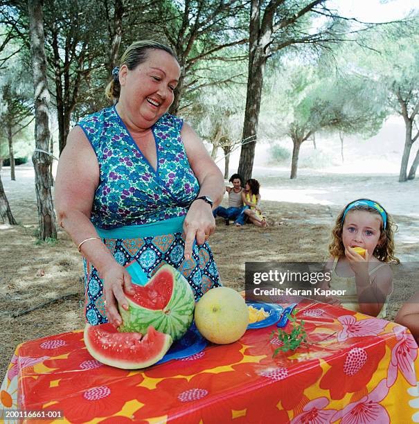 mature woman and girl (2-4) at table outdoors, woman cutting melon - chubby granny foto e immagini stock