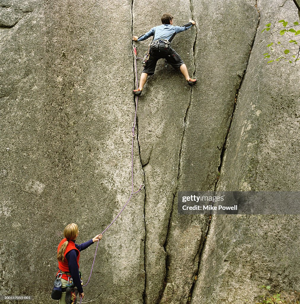 Woman holding rope, looking upward at partner climbing rock wall