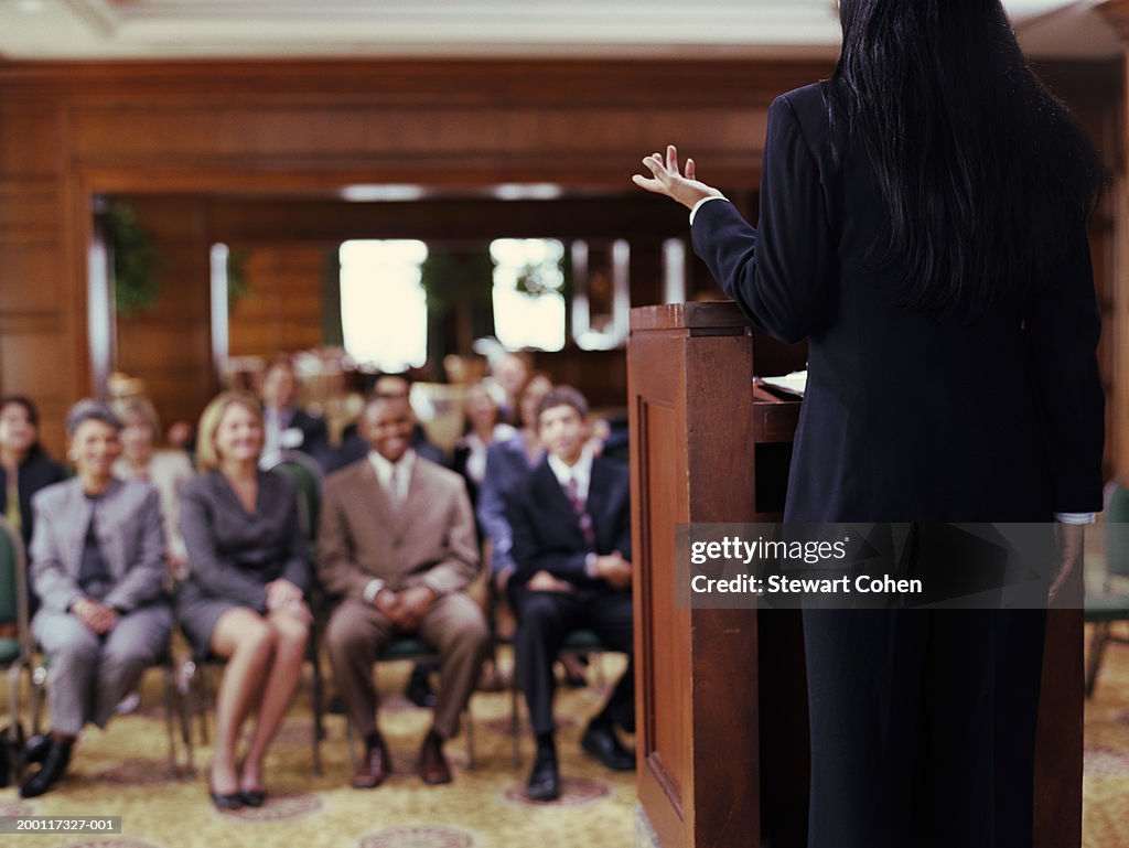 Business woman standing behind podium, speaking to audience, rear view