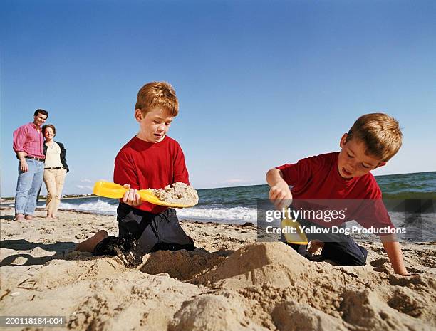 twin boys (6-8) playing in sand on beach, parents watching and smiling - twin boys stock pictures, royalty-free photos & images