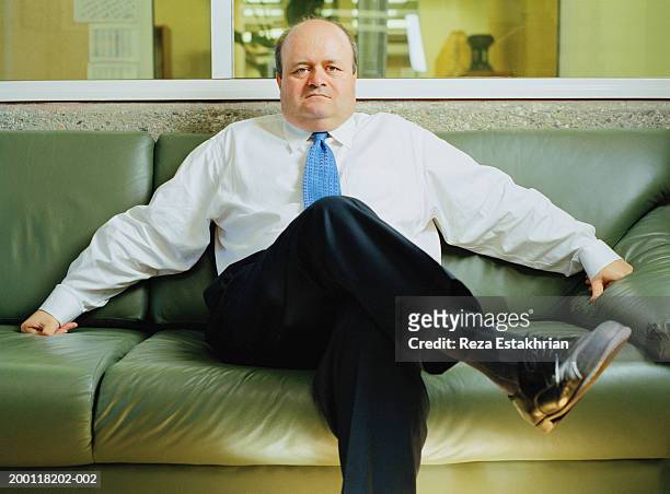mature businessman sitting on leather sofa, portrait - benen over elkaar geslagen stockfoto's en -beelden