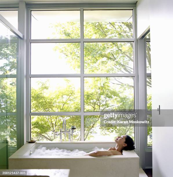 woman soaking in bubble bath, surrounded by glass windows, side view - domestic bathroom stock pictures, royalty-free photos & images