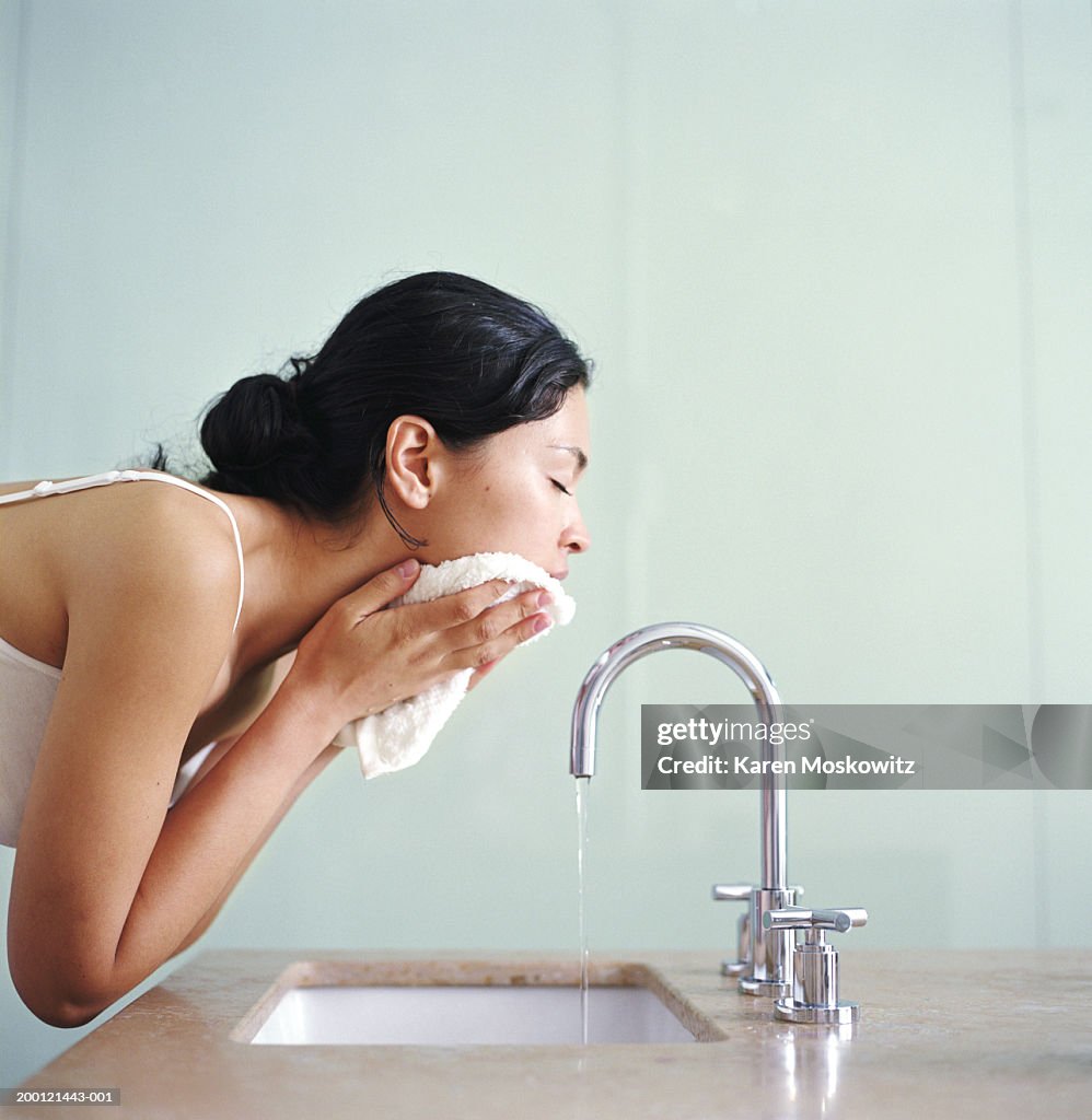 Woman washing face with wash cloth over sink, side view