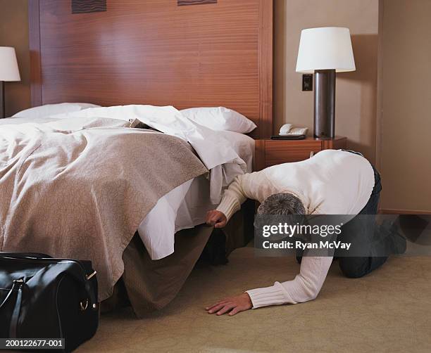 man kneeling on floor in hotel room, looking under bed - paranoia stock-fotos und bilder