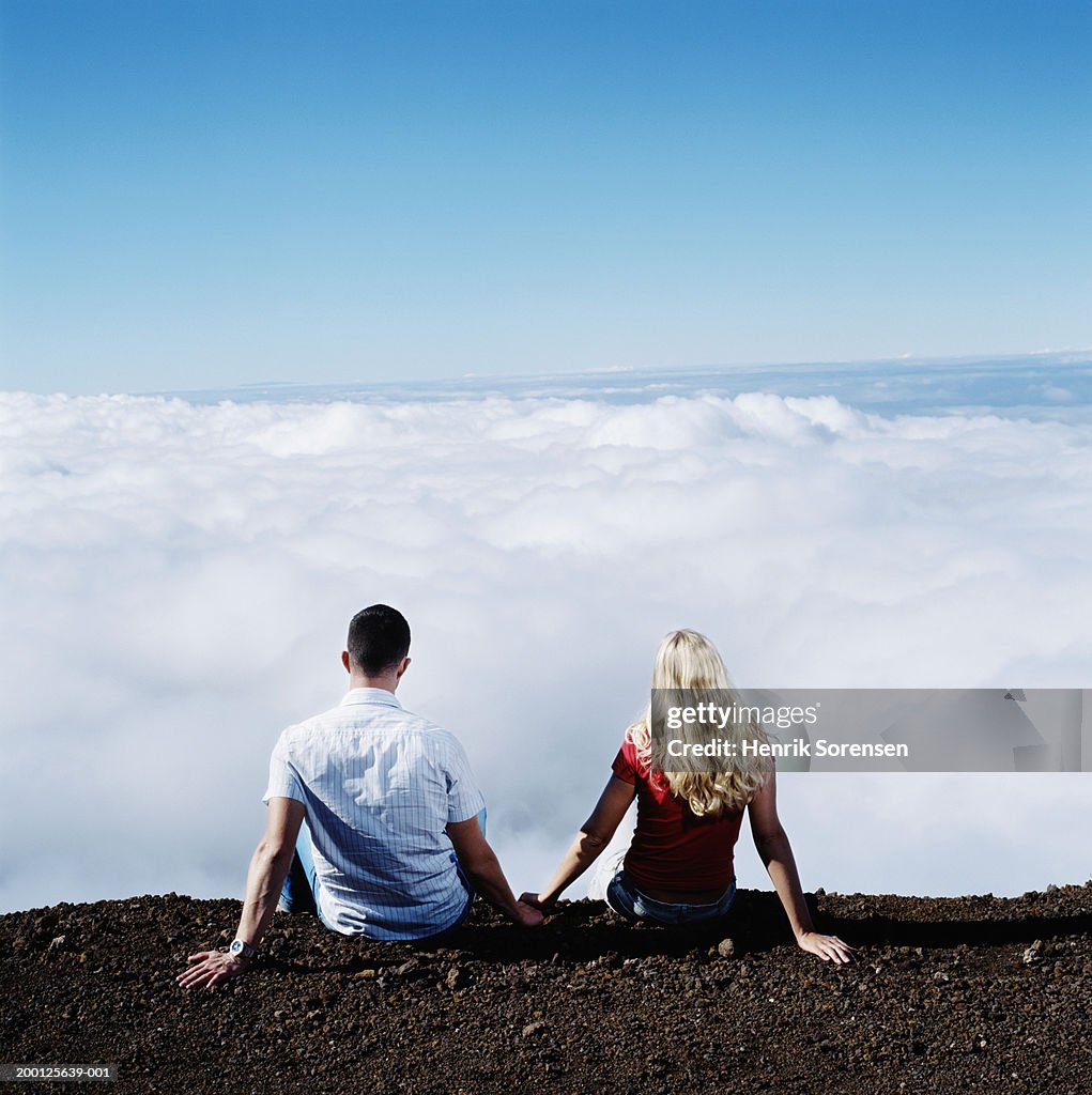 Young couple sitting on mountainside looking over clouds, rear view