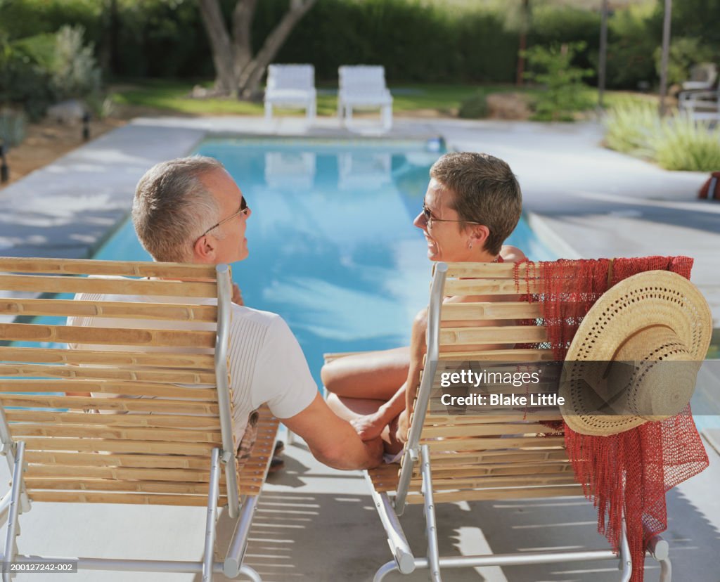 Mature couple sitting by poolside, straw hat hanging from chair