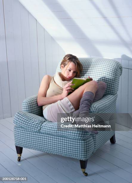 young woman curled up in chair reading book - curled up reading stock pictures, royalty-free photos & images