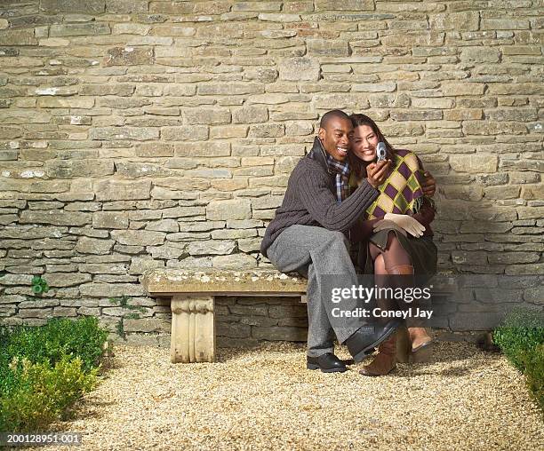 couple on stone bench in front of brick wall, looking at mobile phone - feature phone stockfoto's en -beelden